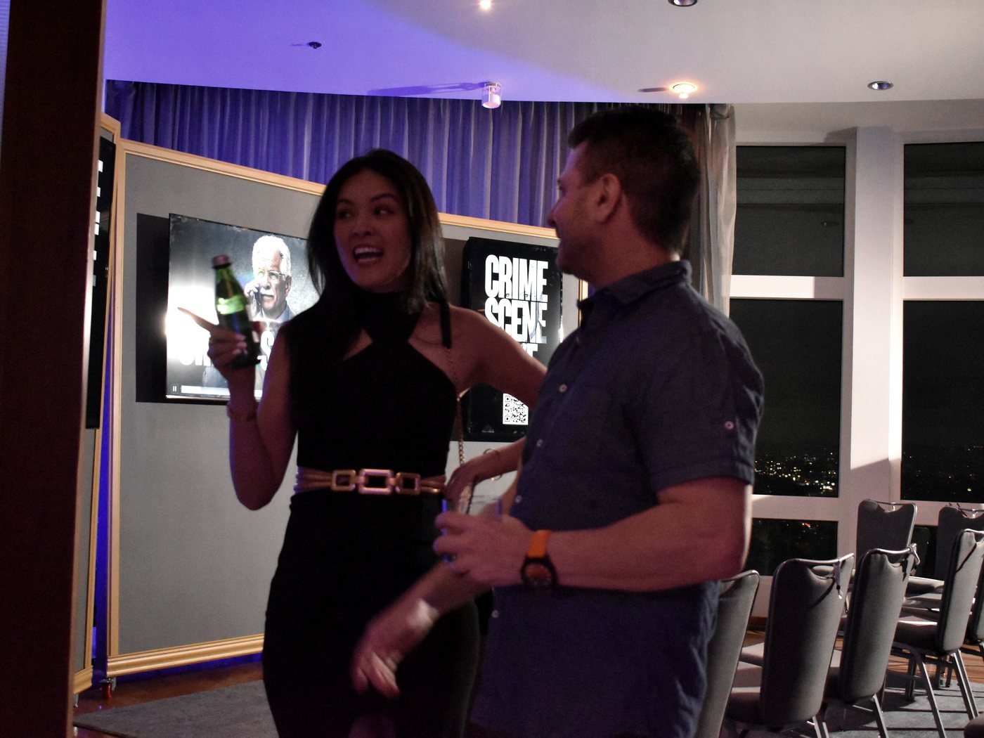 A guest laughing and posing for a photo, drink in hand, with the Crime Scene Live screen behind her